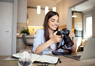 Happy,Young,Photographer,Holding,A,Dslr,Camera,In,Her,Home Happy young photographer holding a dslr camera in her home office. Female photographer smiling cheerfully while working at her desk. Creative female freelancer working on a new project.