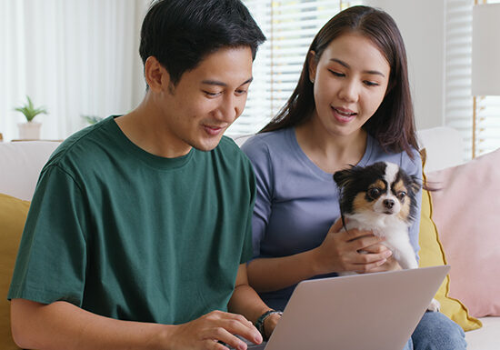 Asia people young family man woman smile happy sit at home sofa couch pay for hotel booking, search choose, buy ticket on laptop social media app travel trip getaway plan tour with cute small pet dog.